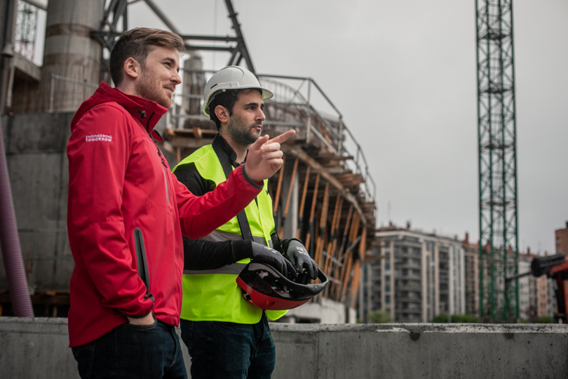 Two men, one in a Danfoss jacket and the other in a safety vest and hard hat, pointing to something on a construction site