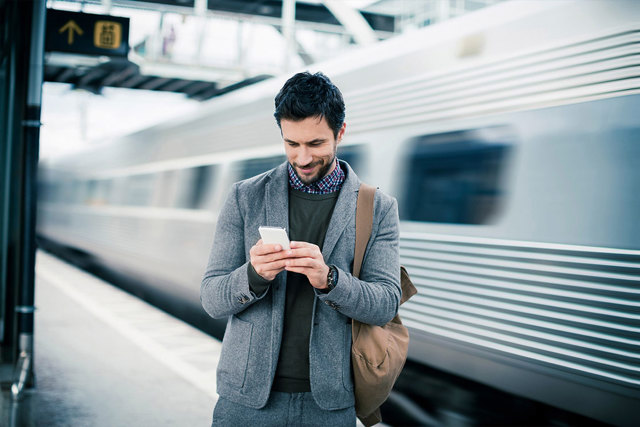 A mid 30's, brown haired, bearded man is at the train station on his smartphone dressed up in a sweater, polo and blazer