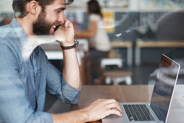 A bearded, hipster-looking gentleman working on a laptop, while talking on the phone at a cafe, clearly looking up Danfoss contact information and calling his local Danfoss office