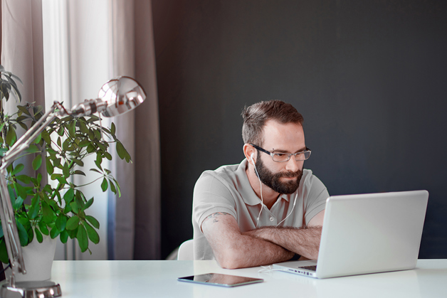 A brown haired, bearded man in a grey polo looking at a laptop at his desk inside a contemporary designed home. 