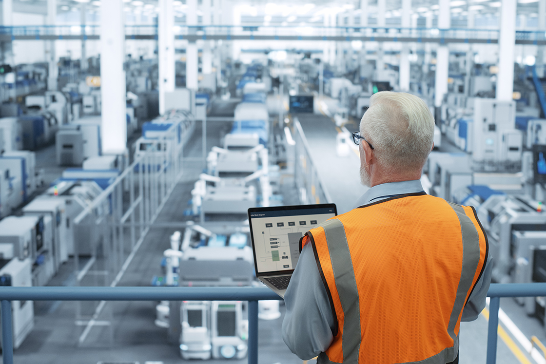 Man in a safety vest looking at his open laptop while standing above a factory floor